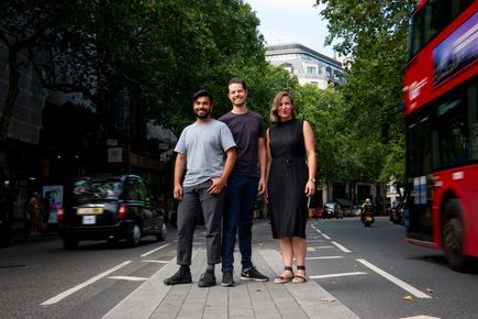 Two men and a woman standing on a central reservation of a road in London. There is a black cab to their left and a red bus to their right