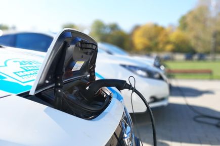 Close up of plug connected to front of electric car. Car is outside against a background of autumnal trees with blue sky above