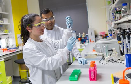 Photograph of female student in a lab with male academic. Both wearing PPE. Student is injecting a liquid into a vial. 