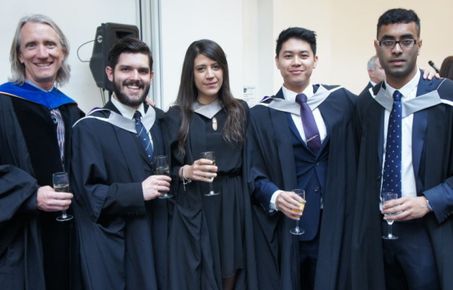 Students standing with a professor wearing graduation gowns and holding glasses of wine