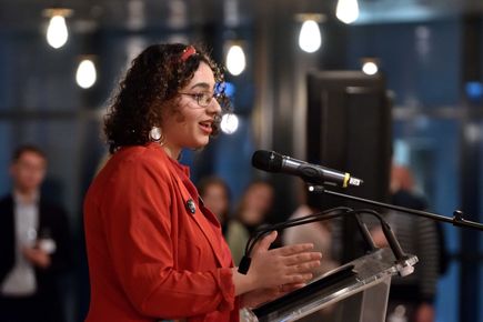 A student, Yasmin Baker, makes a speech at a microphone in a room of people