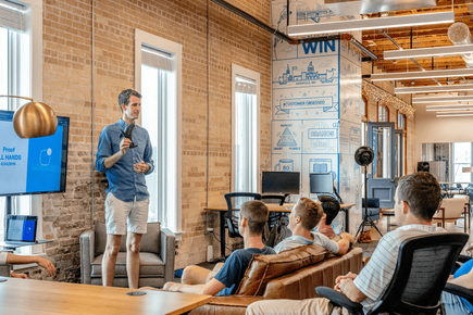 A man giving a talk to four people on a sofa in an open plan, exposed brick office space