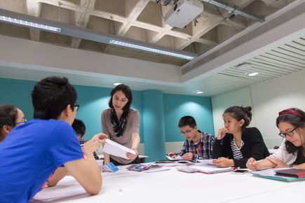 A group of students sitting around a desk in an academic English class talk to a teacher