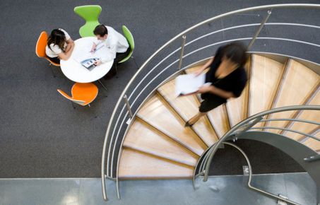 Lady walking down spiral stairs with students sat at the bottom of the stairs working collaboratively 