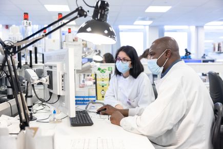 Two researchers wearing lab coats and facemasks work on a computer