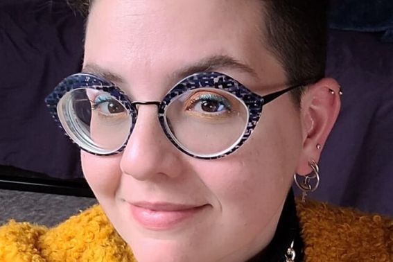 A white non-binary person smiles at the camera with a yellow sweater and big black oval glasses