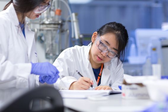 Two women wearing lab coats in a lab environment, one is writing in a notebook