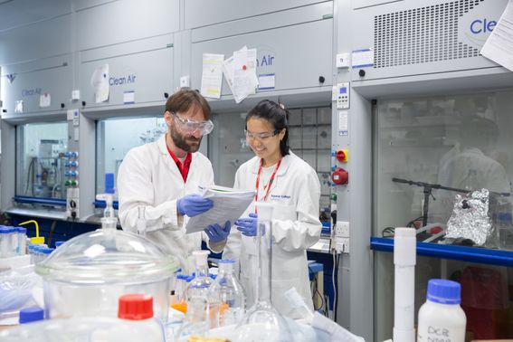 A man and a woman standing in a lab, wearing lab coats and examining a piece of paper held between them