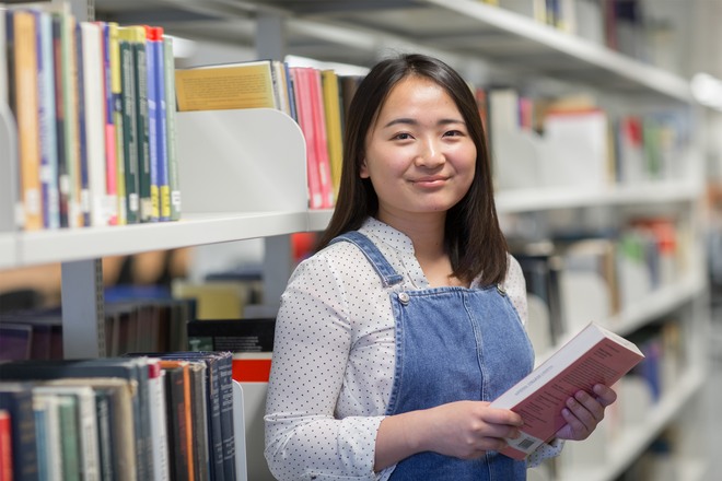 A student smiles at the camera in the library, with a book in hand