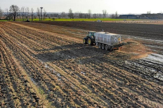 A tractor driving up a field pulling a trailer which is spraying biosolids (manure or treated sewage waste) on a field