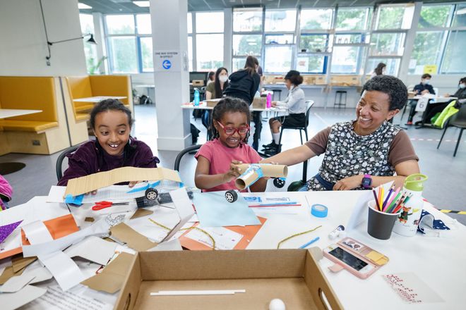 Two children and a woman laugh as they make toy cars from paper and cardboard