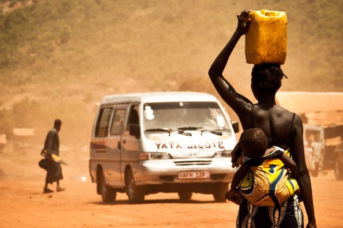 An African woman carrying a water canister on her head and a child on her back, walking along a dusty road with a van coming towards her