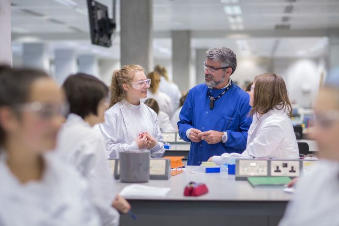 A lecturer in a blue lab coat talks to two students at a lab bench
