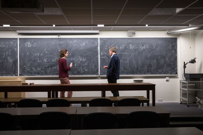 A lecturer and a student discuss equations written on a blackboard