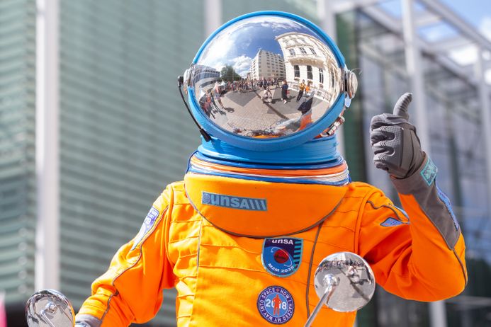 Someone in an orange spacesuit with a reflective globe-shaped helmet gives a thumbs up