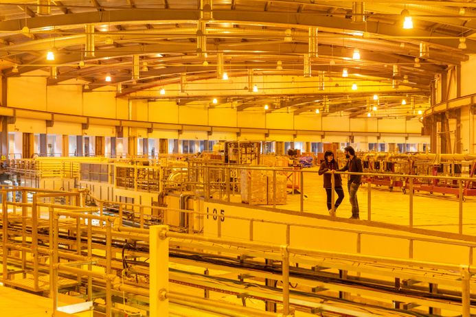 Two people talk in a huge lab that looks like a warehouse with pipes and wires, wholly lit in yellow