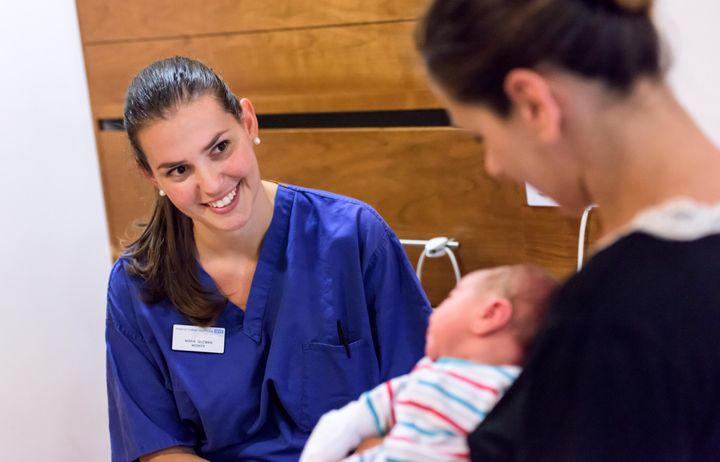 A midwife in blue scrubs smiling at a mother and newborn baby