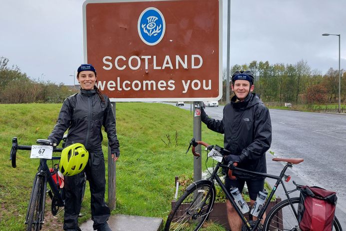 Two cyclists with their bikes, in full rain gear, by a signpost welcoming you to Glasgow