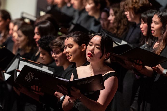 In the foreground, a woman is holding a songbook and singing. In the background you can see other singers in a concert.