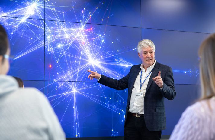 Professor Frank Kelly points at a visualisation displayed on a large screen while talking to a group of students