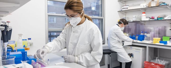 A researcher wearing a lab coat and safety glasses put a vial in a holder in a COVID-19 testing laboratory