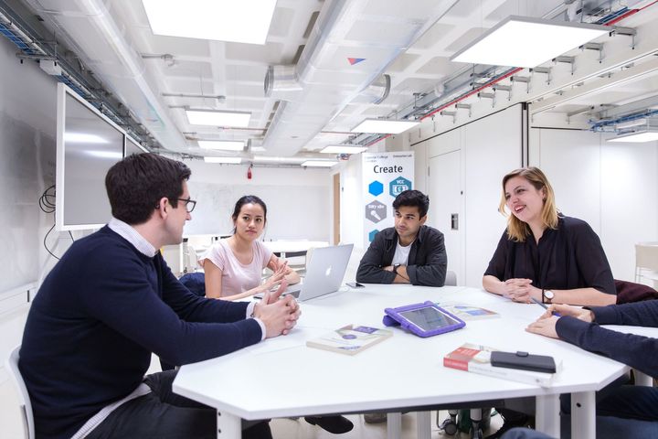 Five students working with laptops and iPads sit around a table