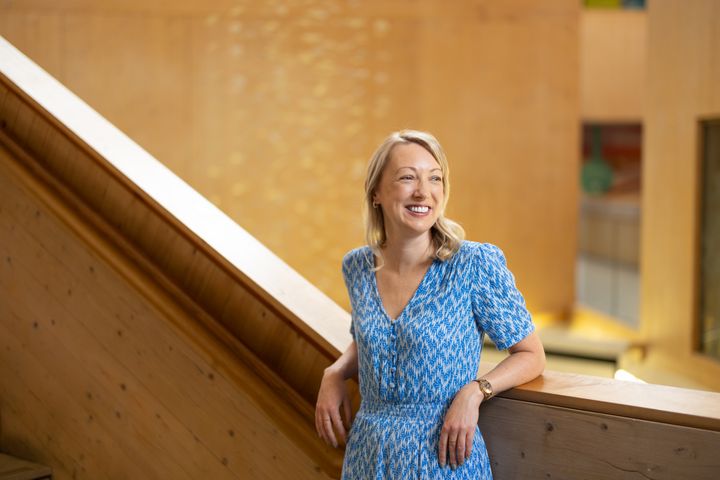 Claire stands against the wooden stairwell smiling, in a blue and white dress.