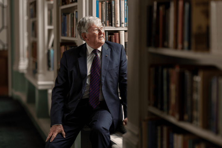 David stands in the library dressed in a suit, surrounded by books, and looks out of the window with a smile on his face.