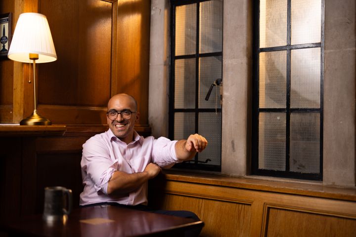 Jad sits in the SU bar, in a warm yellow light, on a table by the windows.