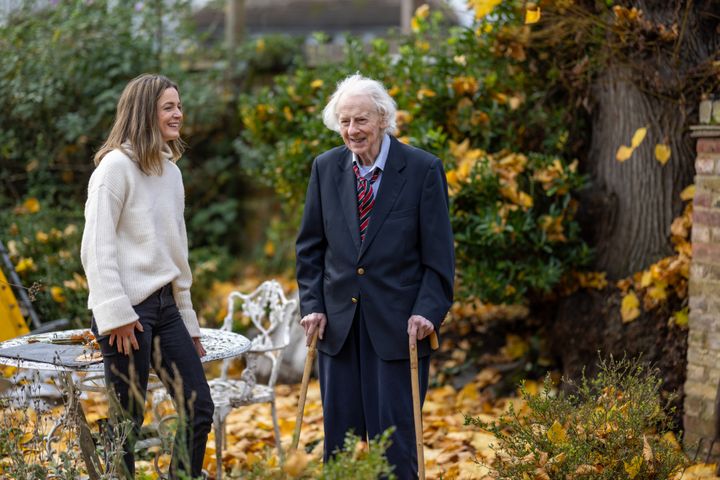 Jenny, wearing a white jumper, stands in a garden on the left next to Sinclair, who is on the right wearing a suit. In the garden, yellow flowers are blooming in the background.