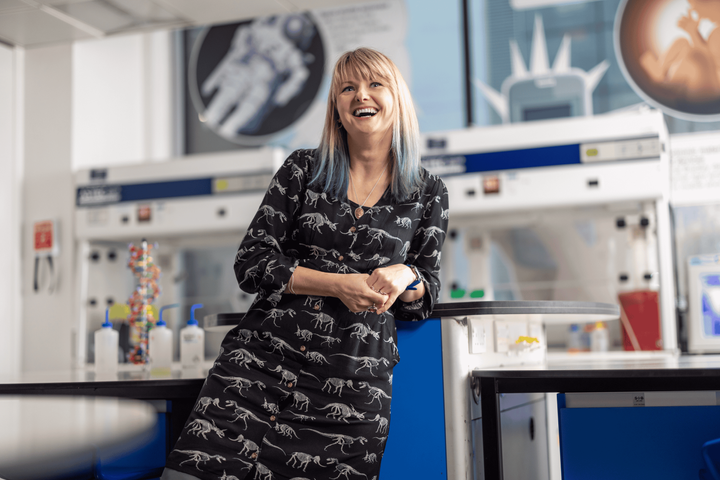 Mel leans against a bench in the Reach Out Lab, wearing a dress with dinosaurs printed on it, laughing into the camera.