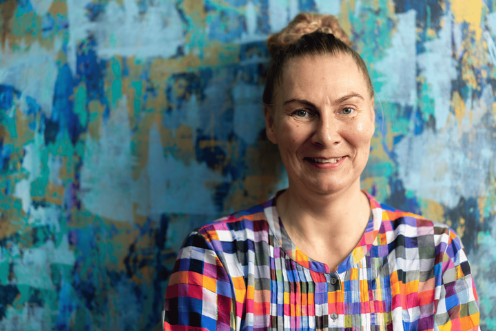 Minna stands against a colourful wall, in a patterned t-shirt, smiling at the camera.