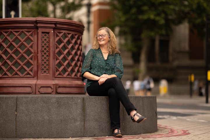 Natasha sits, smiling, on a bench in South Kensington. She is wearing a green shirt and blue glasses.