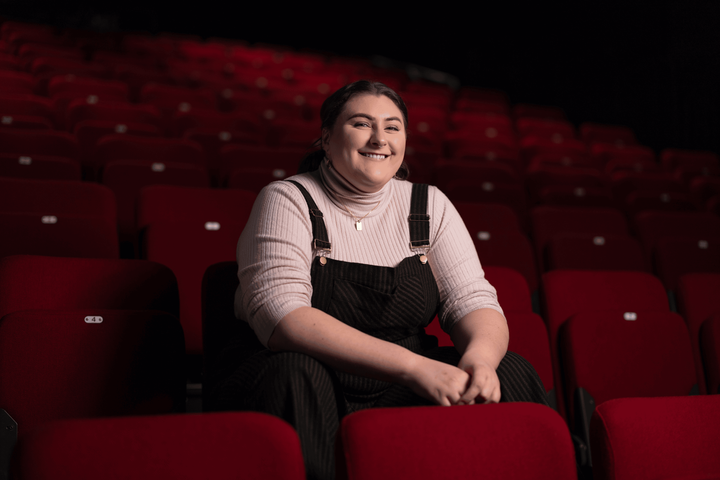 Emma sits in an empty theatre surrounded by red chairs, wearing a white turtleneck and black dungarees, smiling at the camera.