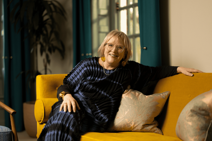 Sara sits, relaxed, on a yellow sofa. She wears a blue dress, glasses, smiling at the camera.