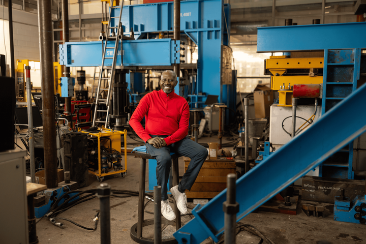 Sunday, dressed in a red sweatshirt and jeans, perches on a stool in an Engineering workspace. He is smiling directly at the camera.