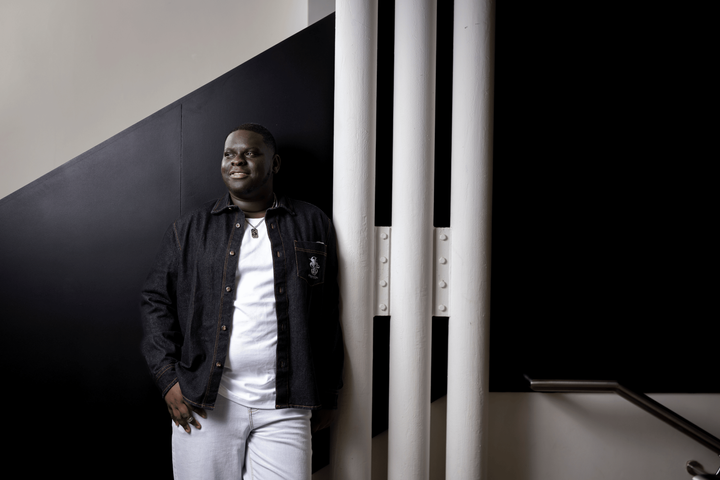 Yemi stands in a stairwell, wearing a denim jacket and white shirt. He is smiling, looking off into the distance.