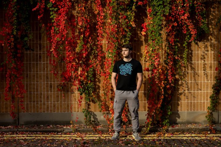Sev stands with his back a wall covered in hanging vines and leaves, in shades of red and green. He has his hands in his pockets, gazing off camera.