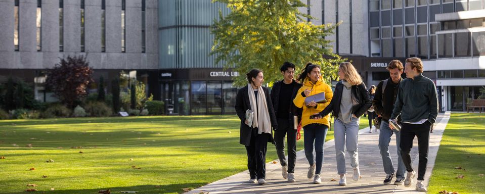 A group of smiling students walk across the Dangoor Plaza, covered in green grass and with the Central Library in the background.