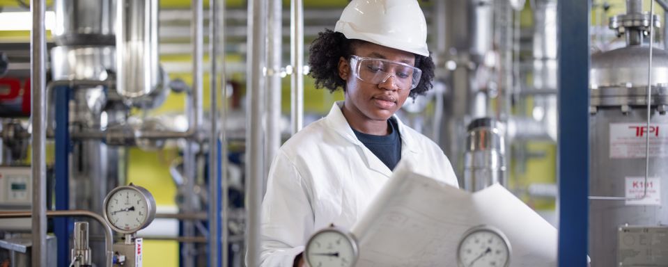 Black woman in a lab coat and hard hat looking at blueprints in the pilot plant