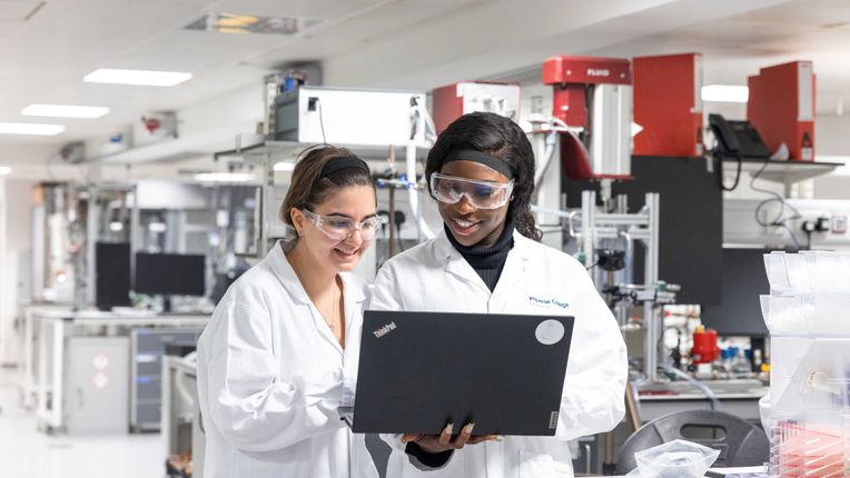 Two students wearing lab coats and goggles in a lab