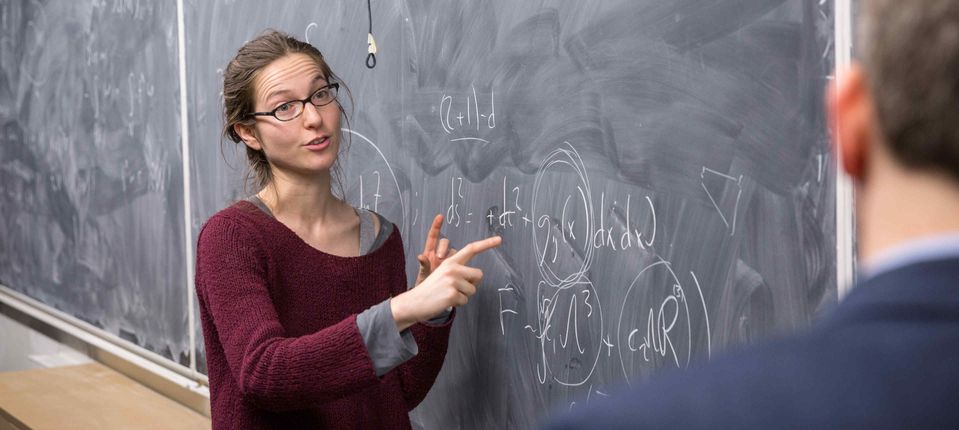 PhD student in the causal set approach to quantum gravity With Professor Toby Wiseman talking in front of a blackboard with equations written up in chalk