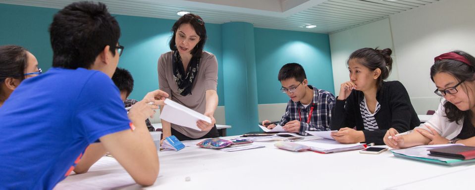 A group of students sitting around a desk in an academic English class talk to a teacher