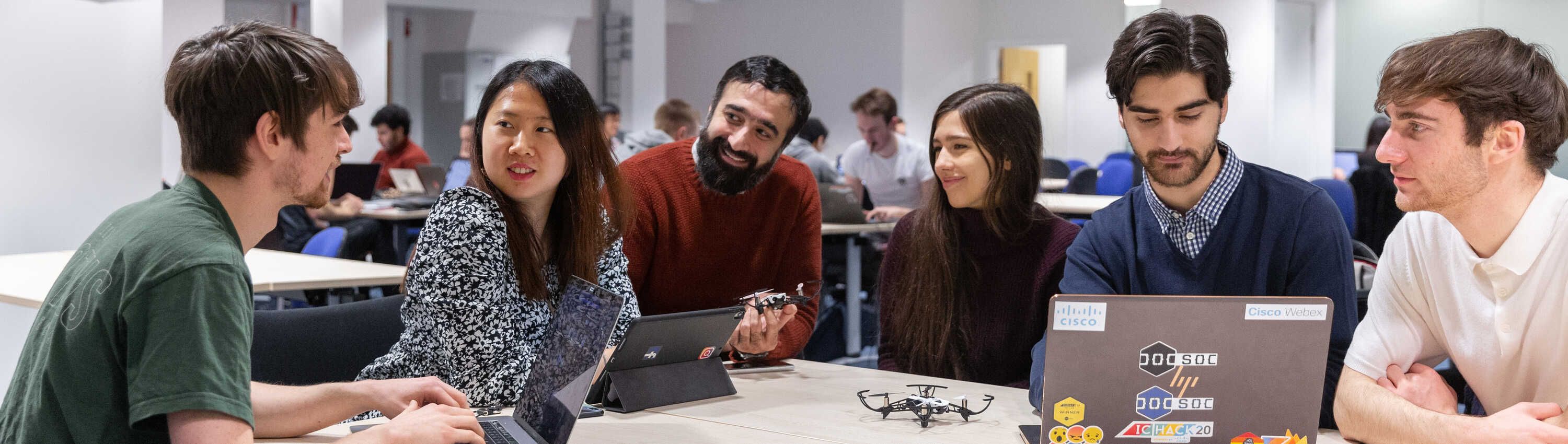 A group of students discussing a research question. A palm-sized quad drone sits on the table in front of them with their laptops and papers.