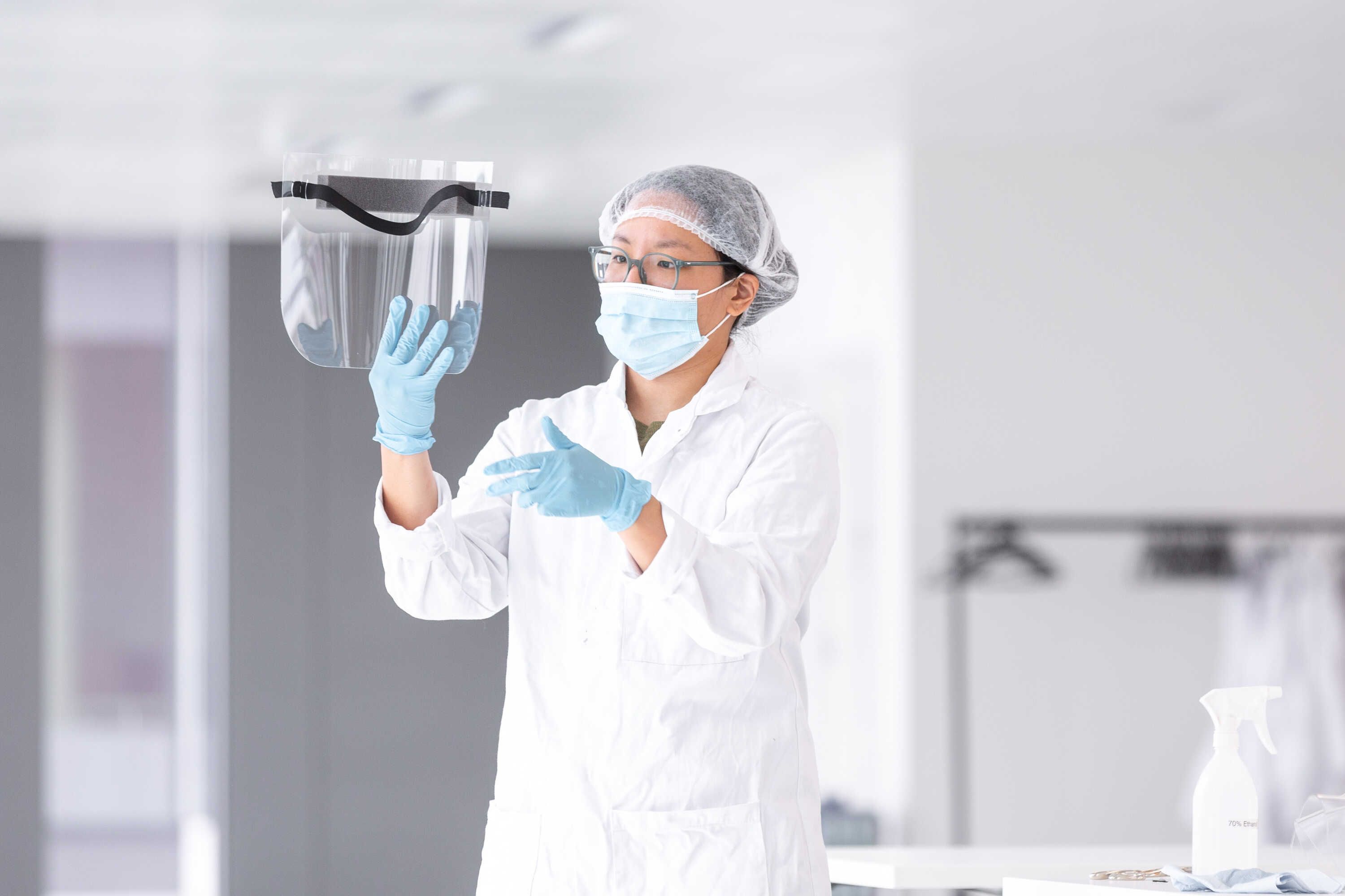A woman in a lab coat and wearing PPE holds up a visor made to protect against COVID-19