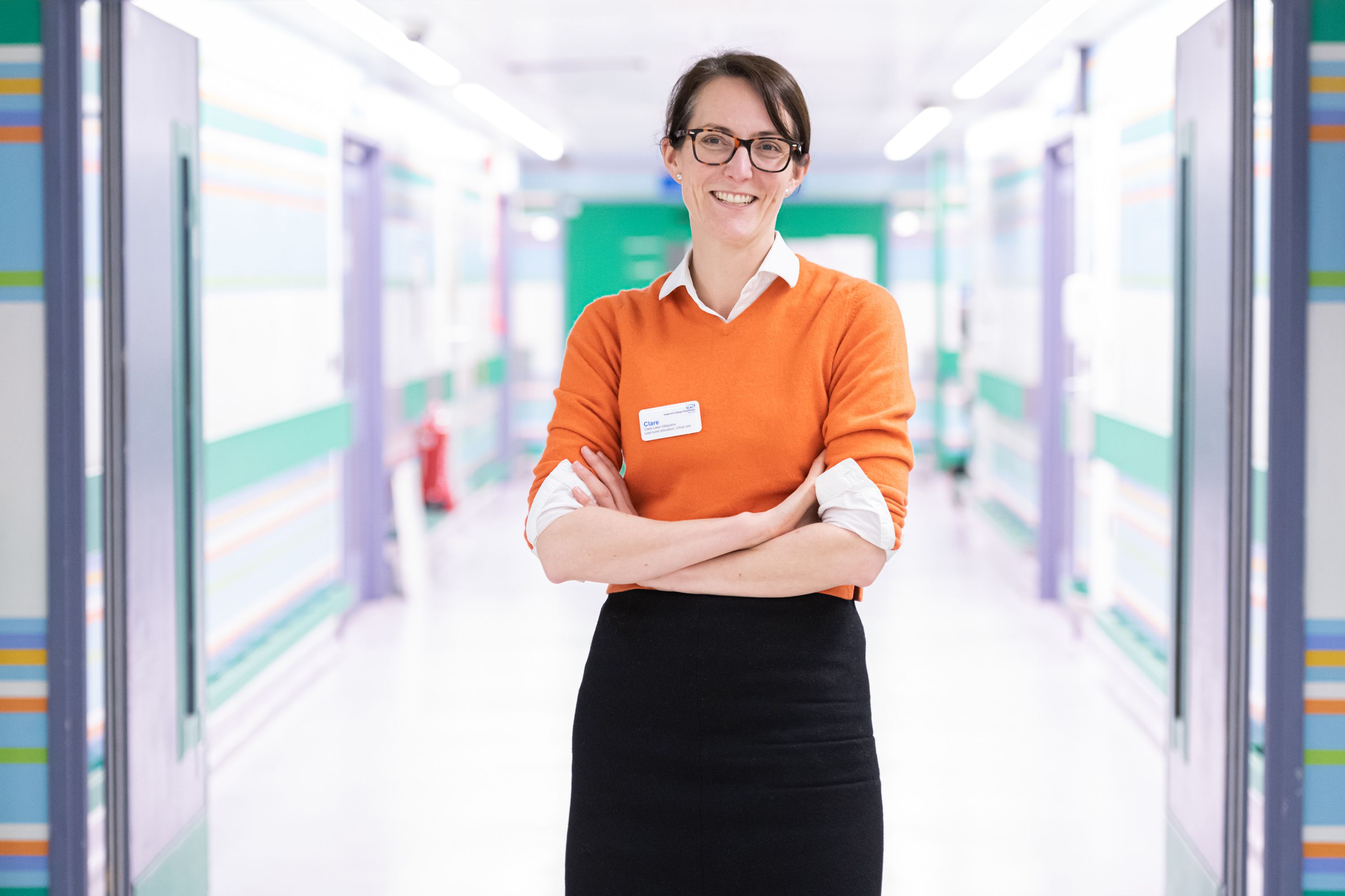 A portrait photograph of Clare Leon-Villapalos smiling as she stands in a white corridor