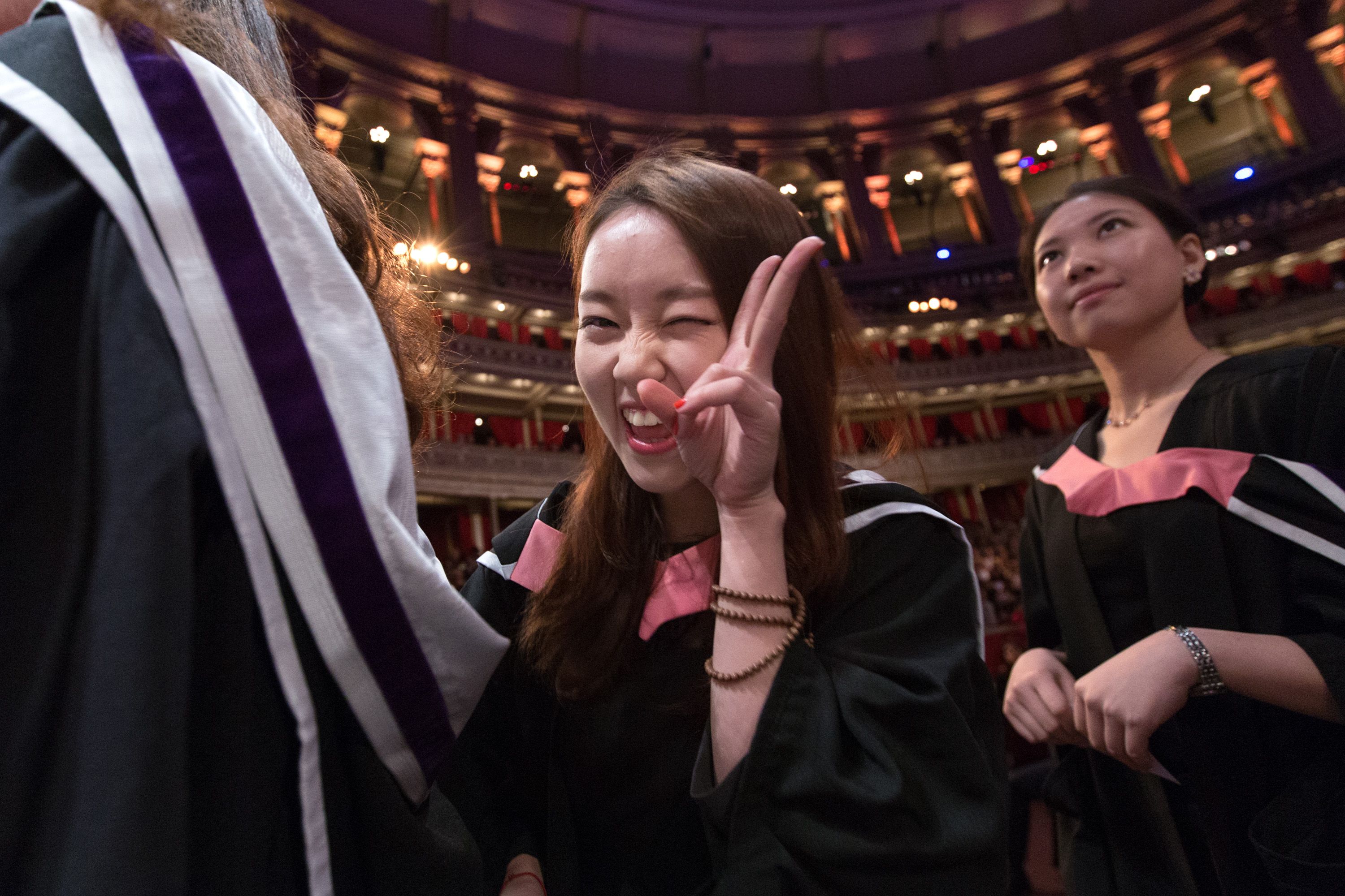 In the Albert Hall, a female student in robes holds her fingers in a peace sign while she queues to graduate