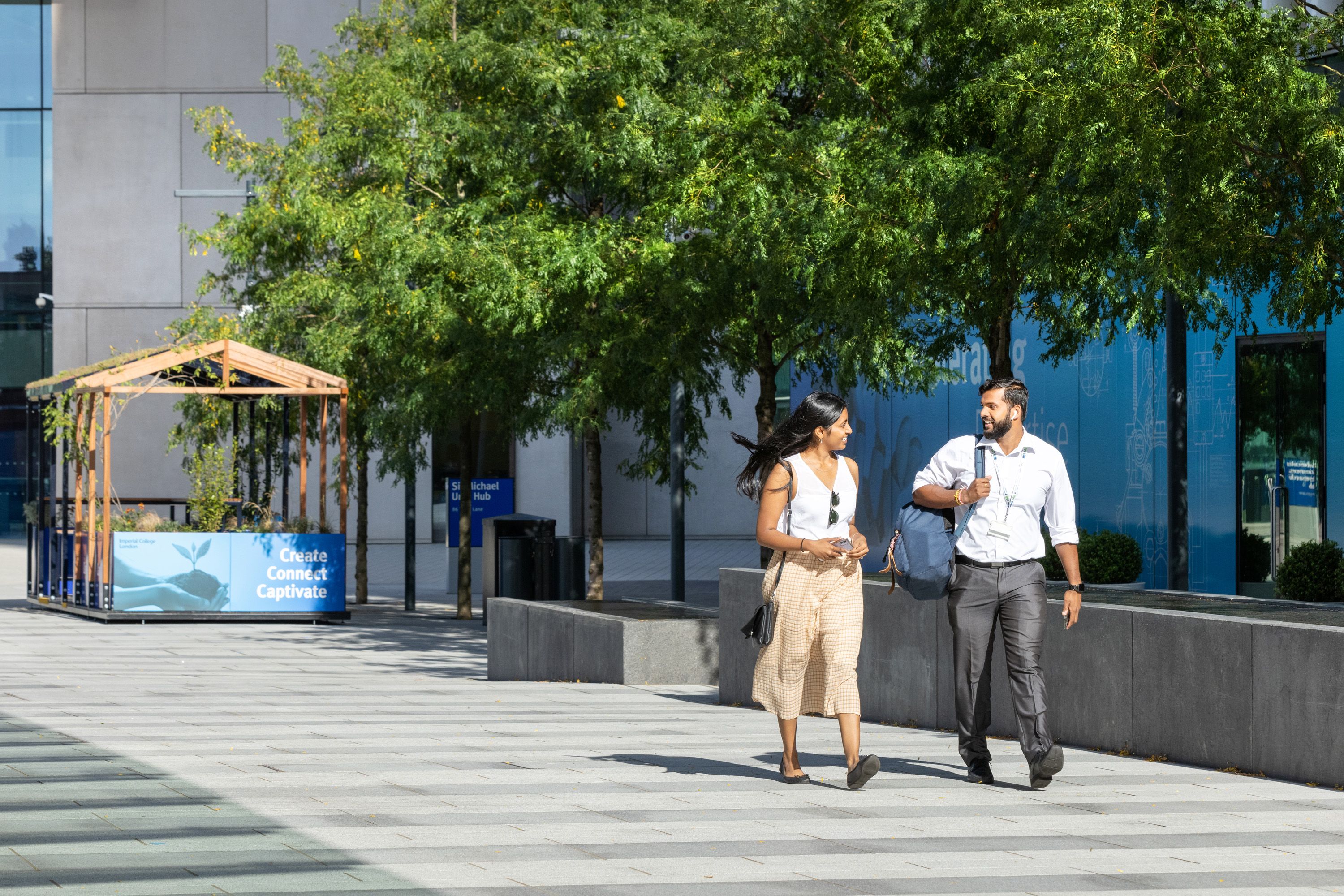 A man and woman smiling and talking walk along a sunny pedestrian area outside the Sir Michael Uren Hub building