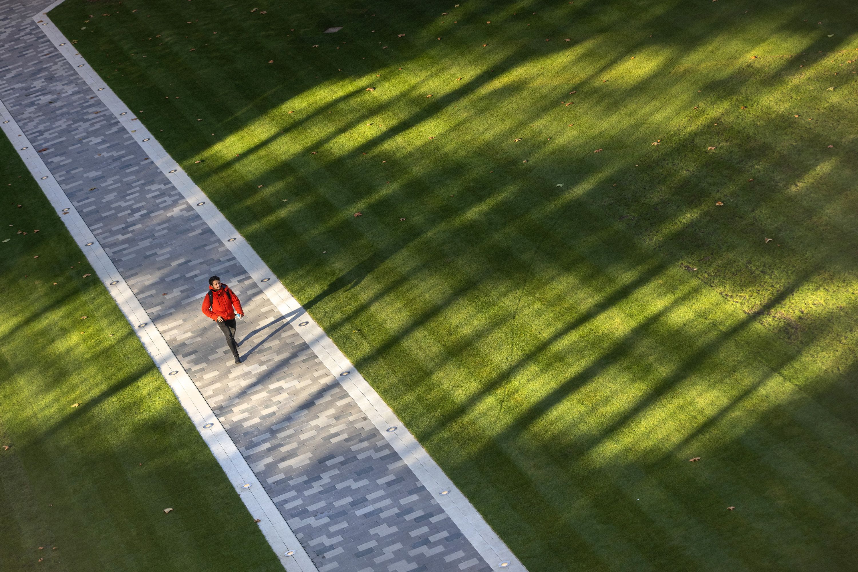 A man in a bright red jacket walks along a paved path that cuts across a grass lawn in a dramatic composition with patterns of reflected sunshine