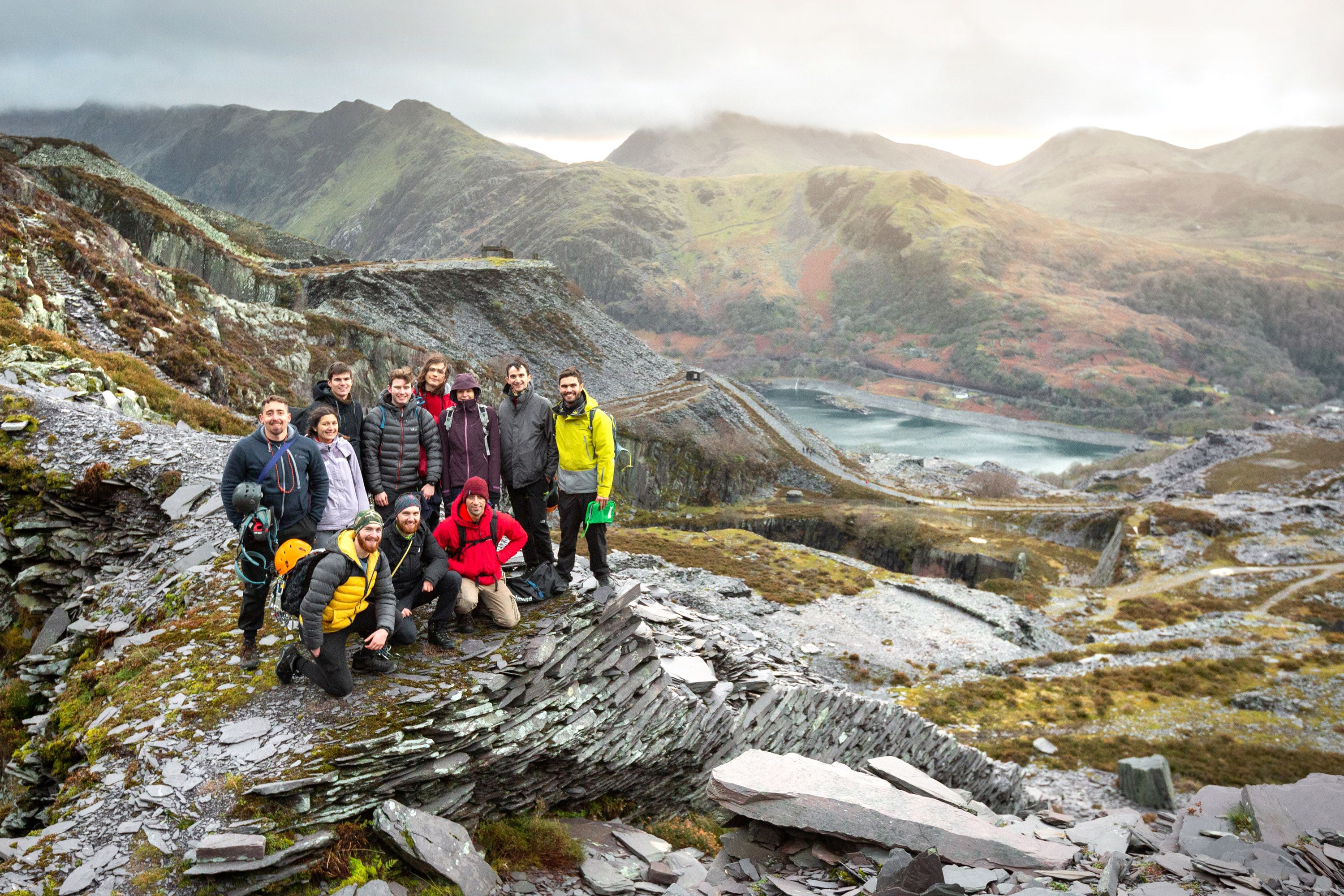 A group of students in weatherproof gear posing for the camera in front of a dramatic background of rocky hills and a lake